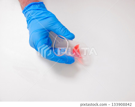 Gloved Hand Holding Cup, Red Droplet, Careful Reagent Transfer On White Surface, Laboratory Preparation 133908842