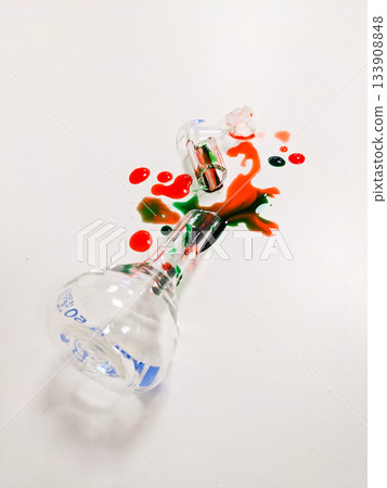 A broken flask and test tube with red and green liquid spills across a clean white background A broken flask and test tube with red and green liquid spills across a clean white background 133908848