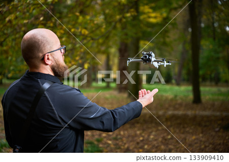 A man with a beard prepares to launch a small drone in a park surrounded by colorful autumn trees. Leaves cover the ground as he focuses on this innovative technology. 133909410