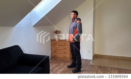 A man in a zipped gray and orange jacket stands confidently with hands in pockets near a window in a modest attic room with wooden furniture and soft daylight A man in a zipped gray and orange jacket stands confidently with hands in pockets near a window in a modest attic room with wooden furniture and soft daylight 133909564