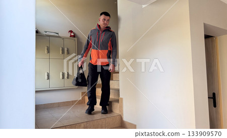A man in a gray and orange jacket walks down tiled stairs in an apartment hallway, carrying a black duffel bag near a wall-mounted locker unit and light-colored walls 133909570