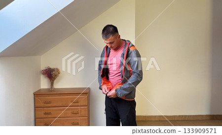 A man in a pink t-shirt and black pants looks down as he zips up a gray and orange jacket in a simple room with wooden furniture and soft natural light 133909571