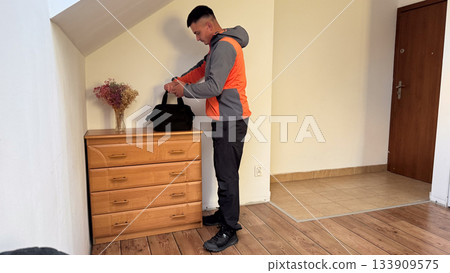 A man in a gray and orange jacket stands by a wooden dresser, securing a black duffel bag with dried flowers in a vase beside him, preparing to leave the room A man in a gray and orange jacket stands by a wooden dresser, securing a black duffel bag with dried flowers in a vase beside him, preparing to leave the room 133909575