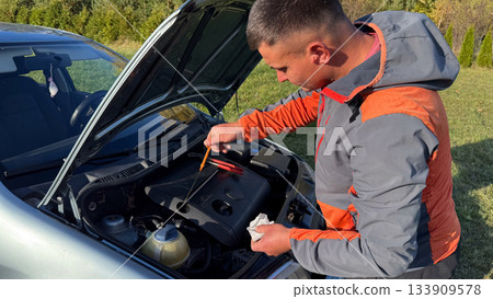 A man in a gray and orange jacket checks the oil level using a dipstick under the open hood of a car during a roadside inspection in a grassy rural area on a sunny day 133909578