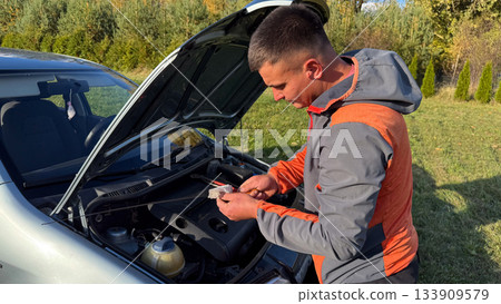 A man in a gray and orange jacket wipes a dipstick with a cloth next to an open car hood, checking engine oil during roadside maintenance in a grassy rural setting 133909579
