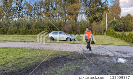 A man in a gray and orange jacket walks across a paved path and grassy area carrying a tool bag, approaching a silver car parked on grass for roadside repair A man in a gray and orange jacket walks across a paved path and grassy area carrying a tool bag, approaching a silver car parked on grass for roadside repair 133909587