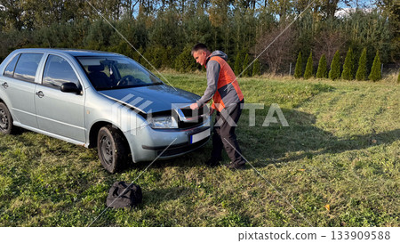 A man in a gray and orange jacket opens the hood of a silver car parked on grass, preparing to inspect and repair the engine during roadside maintenance in a rural area A man in a gray and orange jacket opens the hood of a silver car parked on grass, preparing to inspect and repair the engine during roadside maintenance in a rural area 133909588