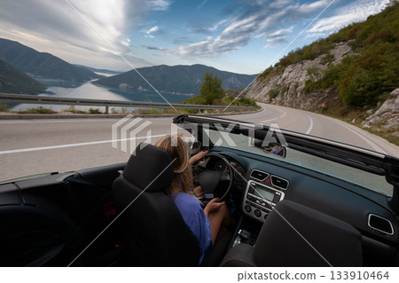 Young woman driving convertible along scenic ocean coast and mountain road at sunset, carefree summer vacation, travel freedom and adventure on coastal highway 133910464
