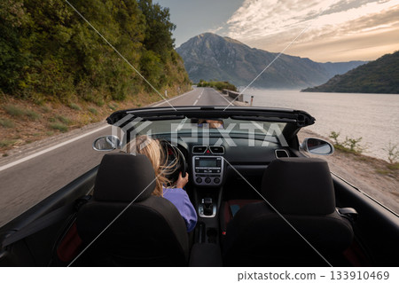 Young woman driving convertible along scenic ocean coast and mountain road at sunset, carefree summer vacation, travel freedom and adventure on coastal highway 133910469