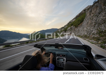 Young woman driving convertible along scenic ocean coast and mountain road at sunset, carefree summer vacation, travel freedom and adventure on coastal highway 133910473