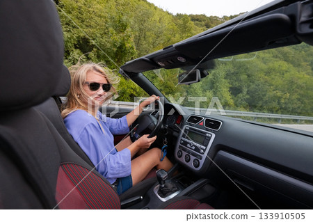 Young woman driving convertible along scenic ocean coast and mountain road at sunset, carefree summer vacation, travel freedom and adventure on coastal highway 133910505