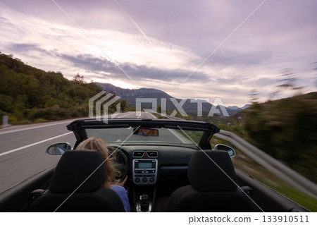 Young woman driving convertible along scenic ocean coast and mountain road at sunset, carefree summer vacation, travel freedom and adventure on coastal highway 133910511