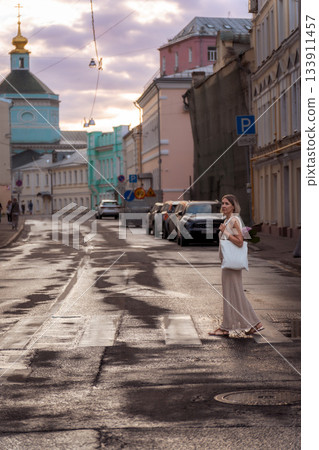 A woman crosses a quiet city street at sunset with a white tote bag and pink hydrangea, warm light reflecting on the wet pavement A woman crosses a quiet city street at sunset with a white tote bag and pink hydrangea, warm light reflecting on the wet pavement 133911457