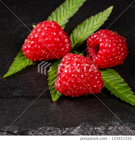 Close-up of three vibrant raspberry berries on a dark stone background 133911583