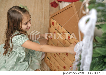 A cute beautiful girl in a green dress opens a cell of the Advent calendar, sitting by the Christmas tree. Looking forward to the New Year and Christmas A cute beautiful girl in a green dress opens a cell of the Advent calendar, sitting by the Christmas tree. Looking forward to the New Year and Christmas 133911710