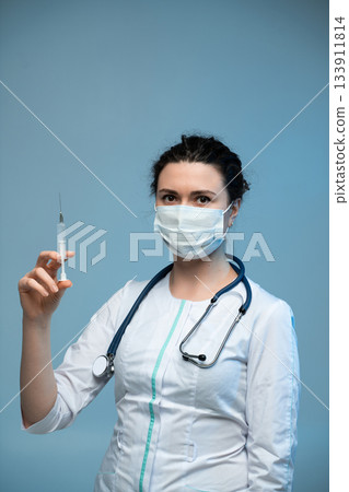 Nurse holding syringe preparing vaccination while wearing medical mask and uniform on blue background Nurse holding syringe preparing vaccination while wearing medical mask and uniform on blue background 133911814