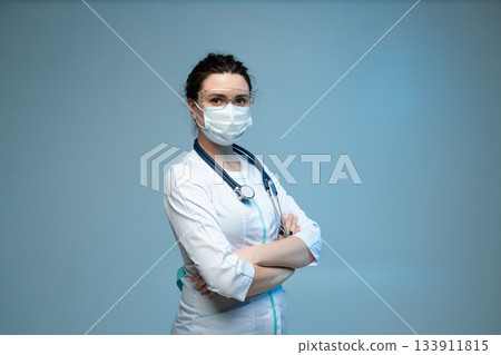 Medical worker in mask and protective glasses standing confidently with folded arms on blue background Medical worker in mask and protective glasses standing confidently with folded arms on blue background 133911815