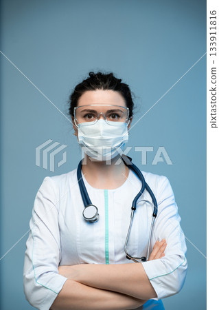 Medical doctor in mask and protective eyewear posing with crossed arms on blue hospital background Medical doctor in mask and protective eyewear posing with crossed arms on blue hospital background 133911816