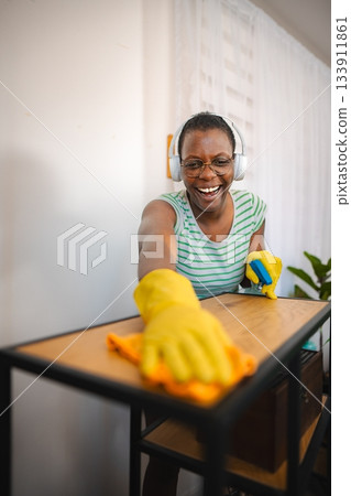 Happy housekeeper wearing headphones and cleaning furniture with detergent and microfiber cloth 133911861