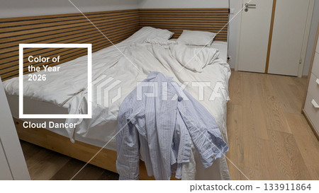 Unmade bed with white bedding in cloud dancer color of the year 2026, blue striped shirt on corner of wooden bed, cozy real bedroom interior with natural wood floor and soft lighting 133911864
