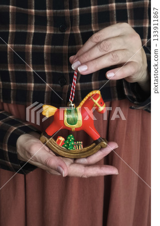 Woman in plaid and terracotta dress holds red rocking horse ornament with tree and gifts, cozy rustic holiday mood 133911867