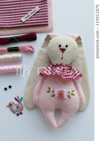 Handmade soft rabbit doll in pastel tones surrounded by pink craft supplies, delicate feminine vibe Handmade soft rabbit doll in pastel tones surrounded by pink craft supplies, delicate feminine vibe 133911870