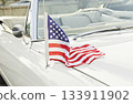 Small American flag mounted on antenna of white vintage convertible, stars and stripes draped over hood in bright daylight 133911902
