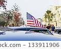 Small American flag on car roof at city street event, people and palm trees in soft background, bright summer vibe 133911906