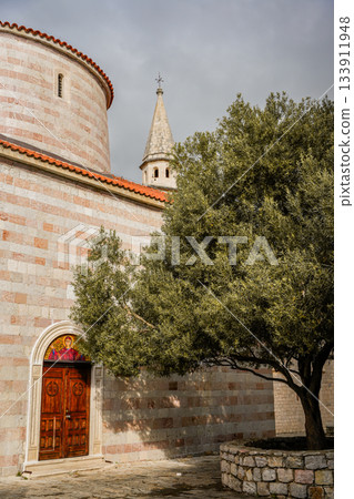 Photo of a large olive tree standing next to a historic church in the Old Town of Budva, Montenegro. Highlights medieval architecture, stone walls, and Mediterranean charm. Ideal for travel, European Photo of a large olive tree standing next to a historic church in the Old Town of Budva, Montenegro. Highlights medieval architecture, stone walls, and Mediterranean charm. Ideal for travel, European 133911948