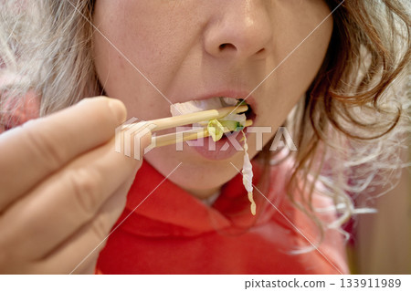 Woman's mouth full of green salad vegetables, using wooden chopsticks for healthy lunch 133911989