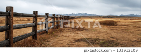 A weathered wooden fence lines the path to a rustic barn on a ranch, surrounded by open fields and distant mountains under gray skies, capturing the peaceful rural atmosphere, banner A weathered wooden fence lines the path to a rustic barn on a ranch, surrounded by open fields and distant mountains under gray skies, capturing the peaceful rural atmosphere, banner 133912109