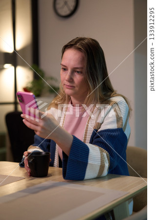 Woman sitting at table looking at smartphone with cup in hand Woman sitting at table looking at smartphone with cup in hand 133912295