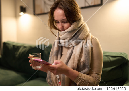 Female with scarf holding water and phone while sitting indoors 133912623