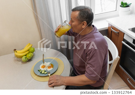 Man drinking fresh juice while having a healthy breakfast at home. Morning routine with nutritious food. Morning Home Cooking Making and Eating Breakfast Man drinking fresh juice while having a healthy breakfast at home. Morning routine with nutritious food. Morning Home Cooking Making and Eating Breakfast 133912764