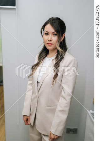 Young Asian woman standing in light business suit with professional demeanor, portraying concept of business professional, medical staff, or successful entrepreneur, looking at camera. Young Asian woman standing in light business suit with professional demeanor, portraying concept of business professional, medical staff, or successful entrepreneur, looking at camera. 133912776