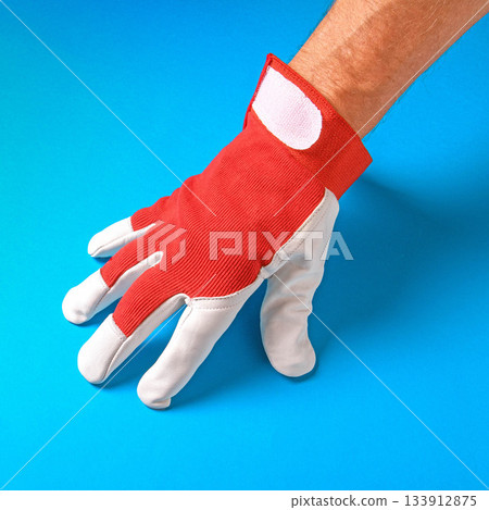 A man's hand wearing a red work glove set against a vibrant blue background 133912875
