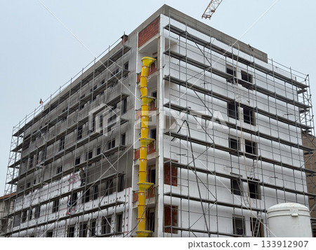 Multi-story building under renovation with scaffolding and chute. City growth, structure, and industrial rhythm in modern architectural process. Multi-story building under renovation with scaffolding and chute. City growth, structure, and industrial rhythm in modern architectural process. 133912907