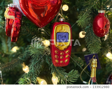 Retro mobile phone ornament with screen icon hanging among Christmas branches. Nostalgic holiday tech memory, playful winter decor and festive tree atmosphere with warm lights. 133912980