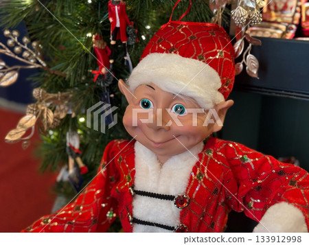 Cheerful elf toy in festive red outfit near Christmas tree. Holiday spirit, winter magic and childhood nostalgia, celebration atmosphere and tradition warmth. Cheerful elf toy in festive red outfit near Christmas tree. Holiday spirit, winter magic and childhood nostalgia, celebration atmosphere and tradition warmth. 133912998