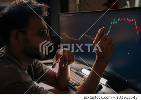Positive young man smiling and pointing at fluctuating financial graph on desktop screen, actively managing cryptocurrency investments and tracking market trends for profitable trading decisions. Positive young man smiling and pointing at fluctuating financial graph on desktop screen, actively managing cryptocurrency investments and tracking market trends for profitable trading decisions. 133913548