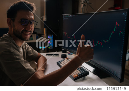 Happy trader male in home office gives thumbs up while viewing rising financial chart on computer screen, celebrating successful online trading, crypto and stock gains. Concept of investing money. 133913549