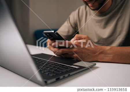 Smiling young businessman working late at night in home office, using smartphone and laptop to manage business tasks and meet deadlines with focused concentration and professionalism. 133913658