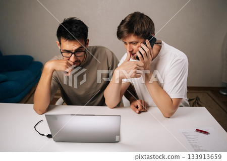 Portrait of two young entrepreneurs in home office collaborate on startup project, one speaks on phone while other concentrates working on laptop, planning and problem-solving. Portrait of two young entrepreneurs in home office collaborate on startup project, one speaks on phone while other concentrates working on laptop, planning and problem-solving. 133913659