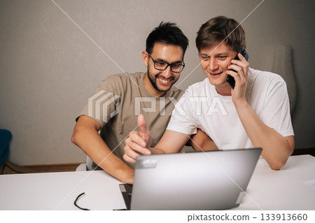 Cheerful successful two young men brainstorming startup ideas, collaborating ona new business project, using laptop and smartphone for communication and strategy in modern home office. Cheerful successful two young men brainstorming startup ideas, collaborating ona new business project, using laptop and smartphone for communication and strategy in modern home office. 133913660
