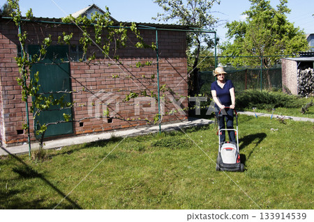 An older woman in a straw hat, working in the spring, mows the grass in her backyard. Concepts of gardening, work, and nature. Concept of movement in adulthood. Lawn maintenance before season. 133914539