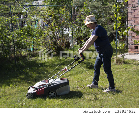 An older woman in straw hat, working in the spring, mows the grass in her backyard. Concepts of gardening, work, and nature. Concept of movement in adulthood. Lawn maintenance before the season. An older woman in straw hat, working in the spring, mows the grass in her backyard. Concepts of gardening, work, and nature. Concept of movement in adulthood. Lawn maintenance before the season. 133914632