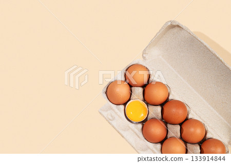 Fresh brown eggs in an open carton with one cracked yolk on a beige background Fresh brown eggs in an open carton with one cracked yolk on a beige background 133914844