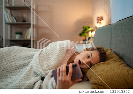 Woman doomscrolling on phone in bed looking shocked at news 133915862
