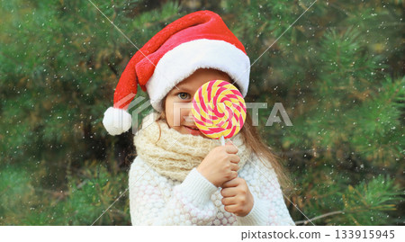 Christmas portrait of happy smiling little girl child in santa red hat with sweet lollipop outdoors 133915945