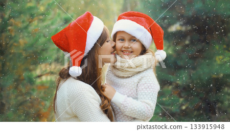 Family, holiday. Happy mother kissing her little girl child in santa hat near Christmas tree outdoor Family, holiday. Happy mother kissing her little girl child in santa hat near Christmas tree outdoor 133915948
